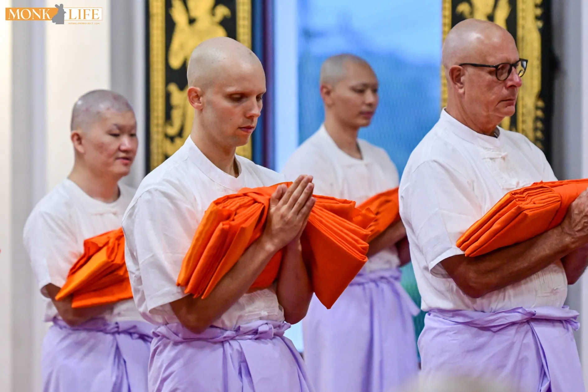 Four ordinands in white robes standing in line with palms together, each holding folded orange robes