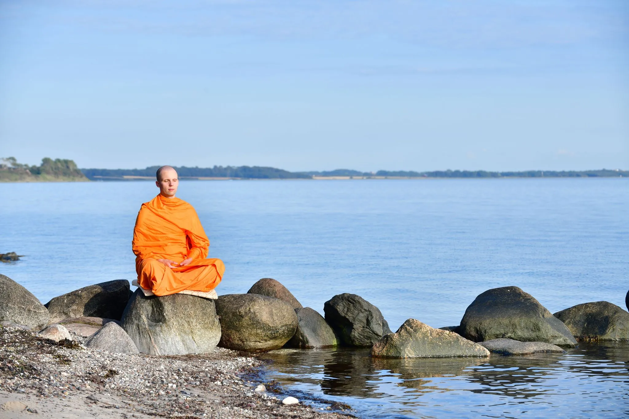 A monk in orange robes meditating cross-legged on a rock at the water's edge