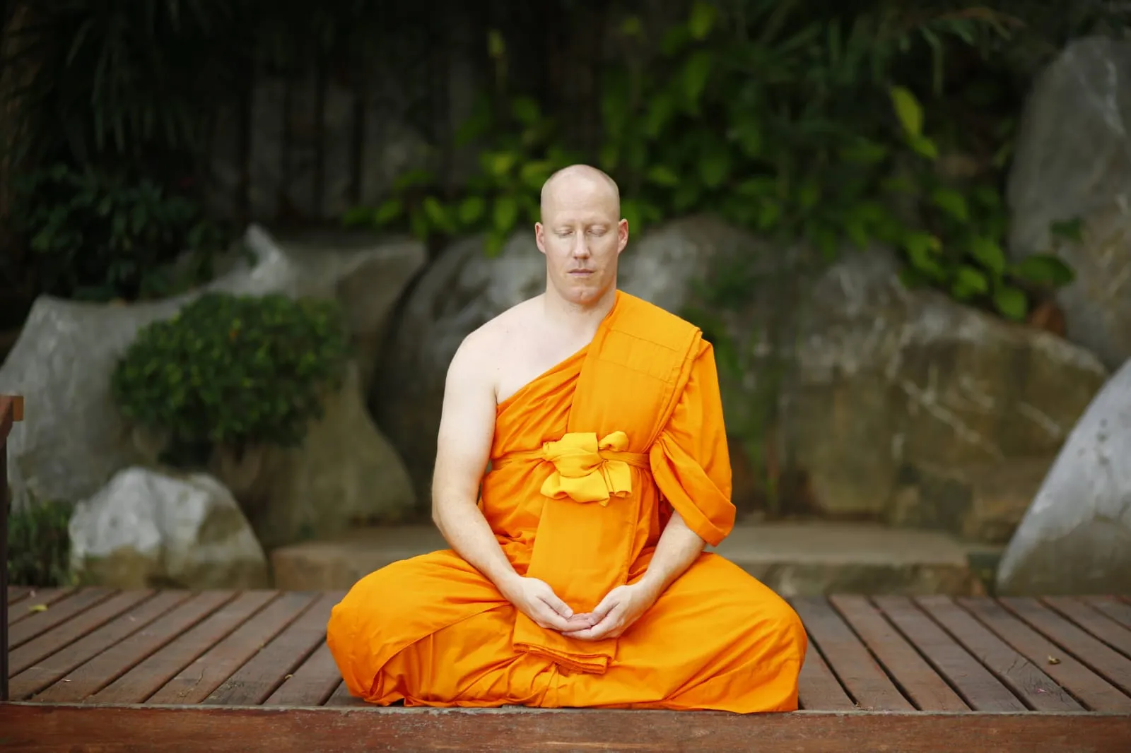 A monk meditating cross-legged on a wooden deck in front of rocks and greenery