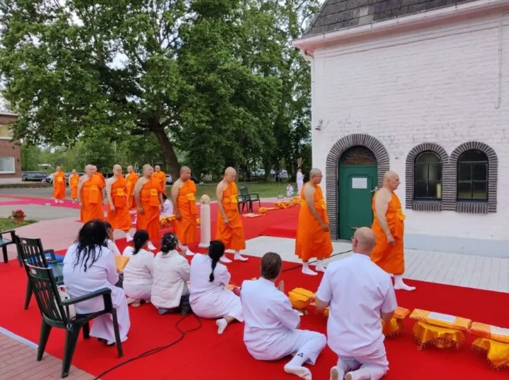 Monks in orange robes processing along a red carpet outside the temple while people in white kneel in front of them