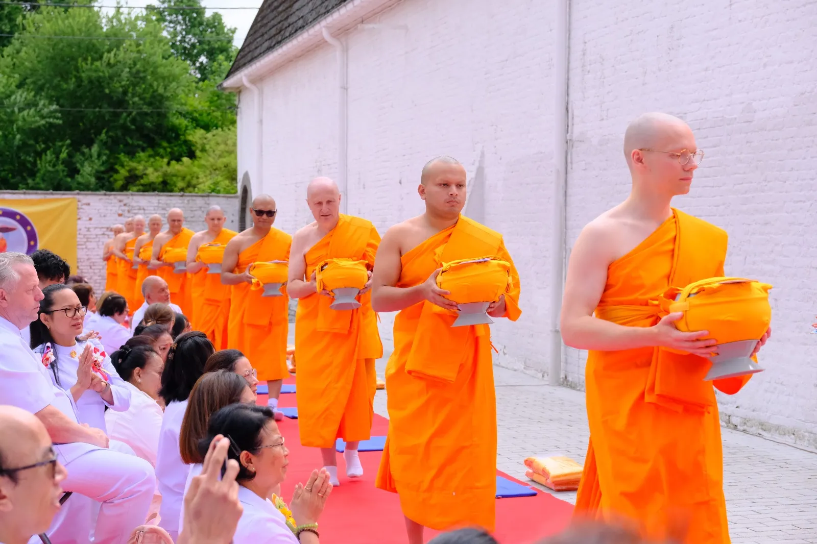 Monks walking in single file holding alms bowls as seated guests greet them with palms together