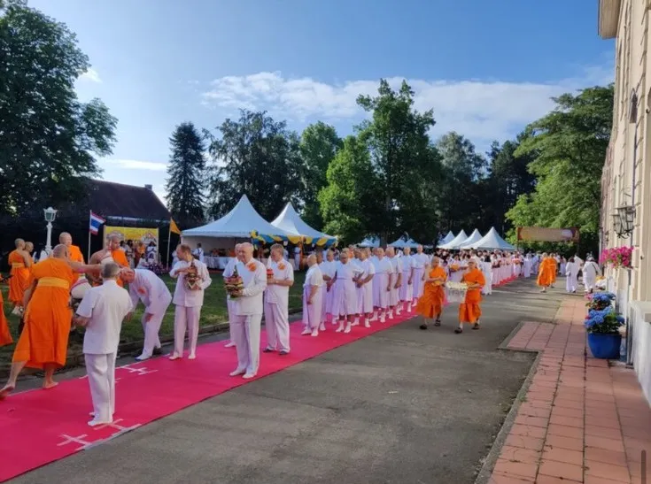 A long line of monks and guests in white walking a pink carpet through the temple grounds past marquees